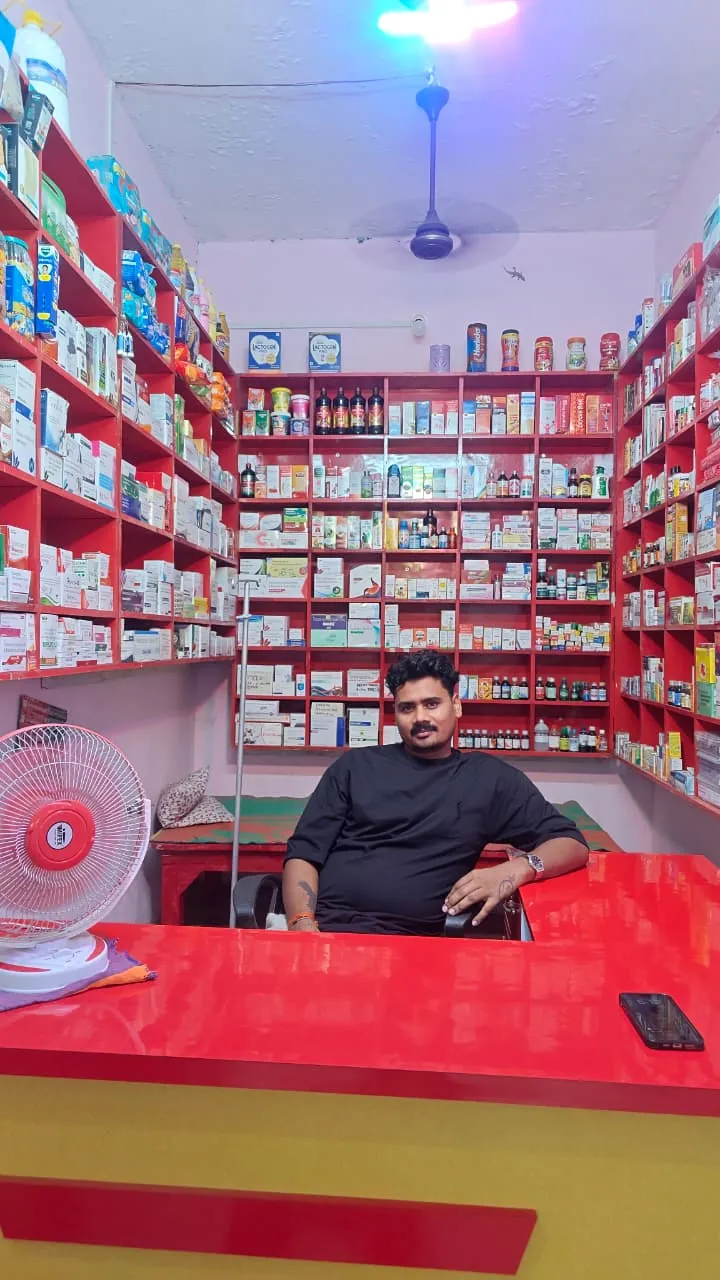 The pharmacist at Sneha Pharmacy's red counter, shelves of medicines behind him